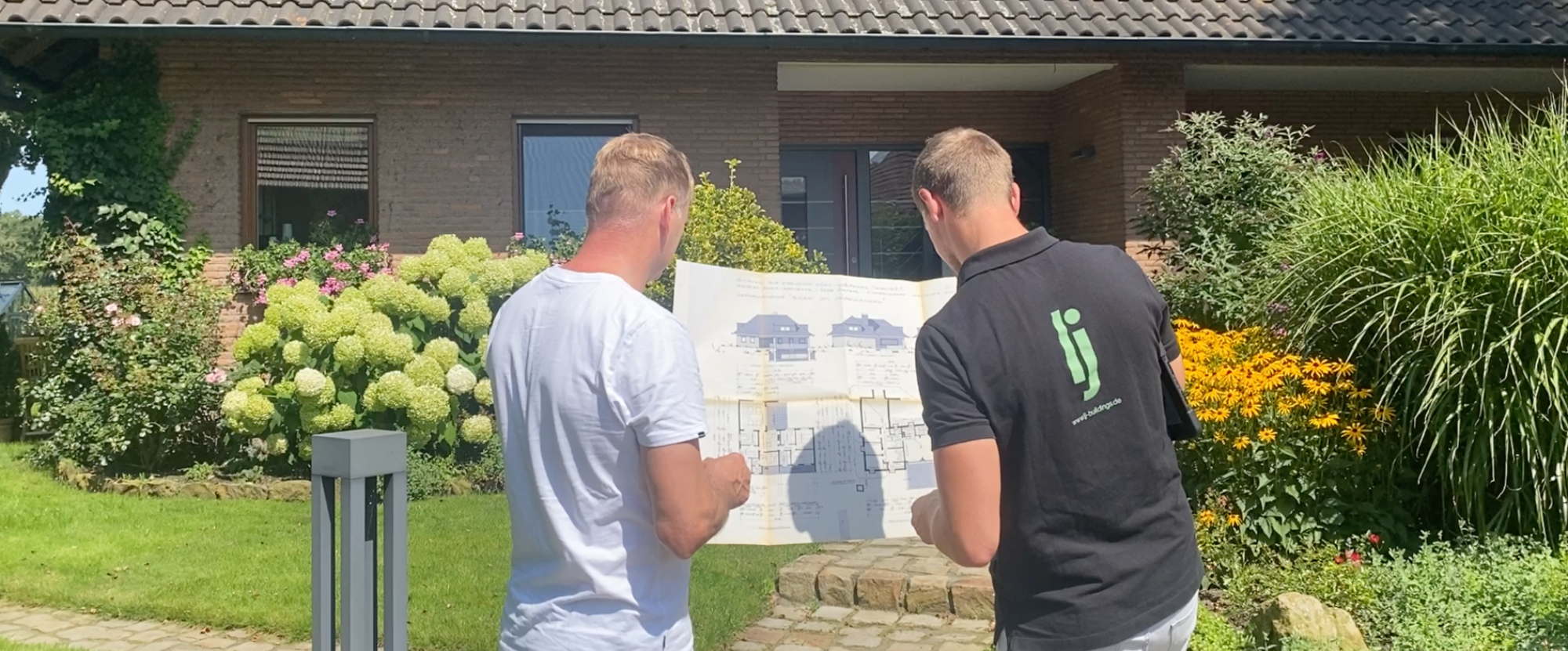 Zwei Personen betrachten einen Plan vor einem Haus mit Blumen im Garten.