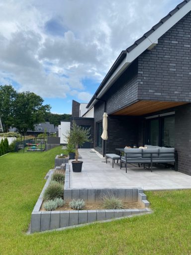 Modernes Haus mit Terrasse und Pflanzbeeten, umgeben von grünem Rasen.