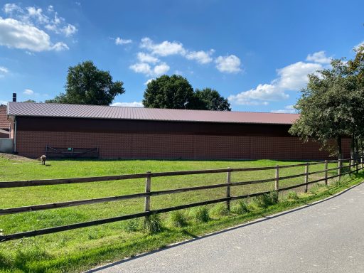 Eine große, braune Scheune umgeben von Wiese und Bäumen unter blauem Himmel.