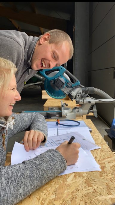 Zwei Personen arbeiten an einem Tisch, während eine Person Notizen macht und lächelt.